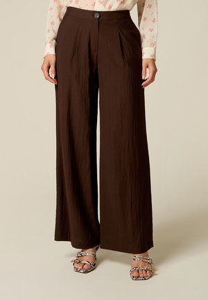 Persoon draagt hoog uitgesneden, wijde donkerbruine broek met knoopsluiting, gecombineerd met een bloemige blouse en sandaaltjes met gestreept patroon.