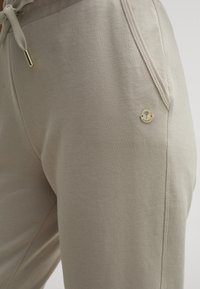 Beige Jogginghosen aus weichem Baumwollmix, mit einem Kordelzugbund, seitlicher Tasche und einem kleinen metallischen Logo-Detail in der Nähe der Tasche.