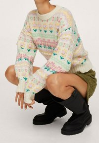 Pull tricoté multicolore avec des motifs floraux et géométriques dans des tons pastel sur un fond blanc cassé, avec des poignets et une base côtelés.