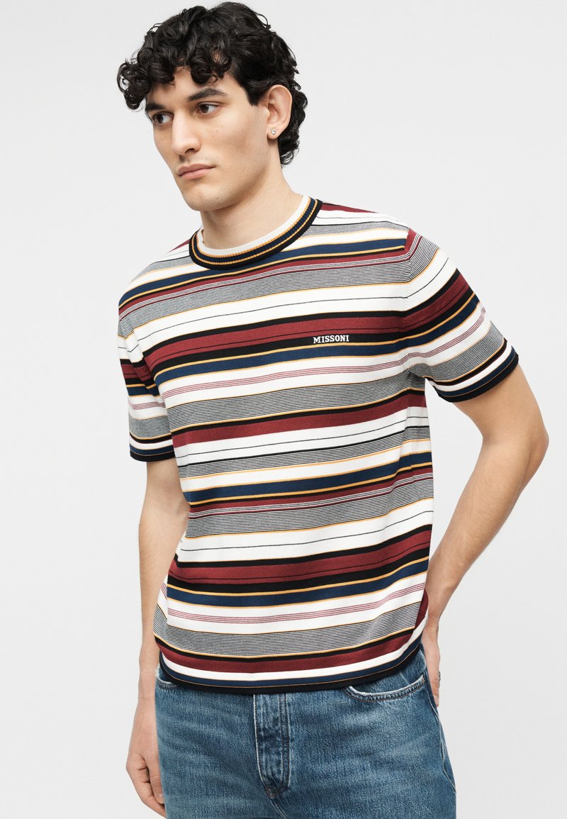 Young man with curly hair wearing a short-sleeve striped T-shirt and jeans, standing with one hand in pocket, looking slightly left.