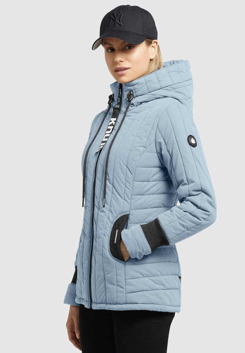 Hellblaue gesteppte Jacke mit Kapuze, schwarzen Akzenten, Reißverschlusstaschen und Kordelzug; kombiniert mit einer dunklen Baseballmütze.