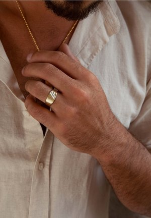 Gouden ring met een geometrisch ontwerp bovenop, gedragen aan een hand met een lichte huid, met kort, donker haar. Linnen shirt zichtbaar.