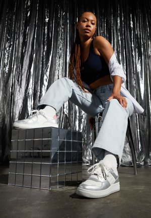 Beli in srebrni športni copati z gladko teksturo, s poudarki v roza barvi, postavljeni na zrcalno kocko, v kombinaciji s svetlo modrimi širokimi hlačami.