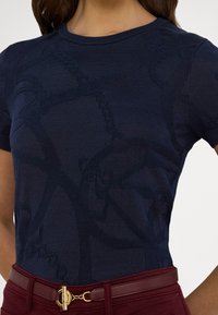 Sötétkék kötött póló texturált absztrakt mintával, kerek nyakkivágással és rövid ujjakkal. Bordó, magas derekú nadrággal és arany csattal párosítva.