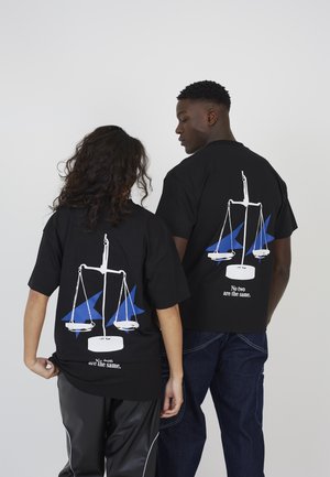 Twee mensen dragen zwarte shirts met witte schubben en blauwe driehoeken, de tekst luidt "Geen twee zijn hetzelfde," staand tegen een effen achtergrond.