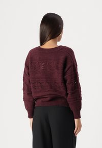 Vinröd stickad tröja med texturerade mönster, ribbad fåll och lös passform. Synligt hålmönster på ryggen, långa ärmar.