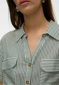 Camicia a righe verdi e bianche con maniche corte, chiusura a bottoni sul davanti, due taschini sul petto e un colletto appuntito. Tessuto di cotone a trama.