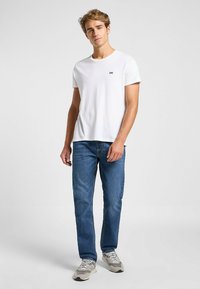Camiseta de algodón blanca con un pequeño logo negro de "Lee", combinada con jeans de denim azul y zapatillas grises, de pie contra un fondo neutro.