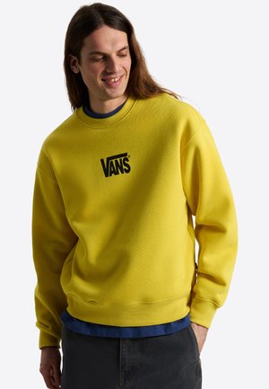 Mand med langt brunt hår iført en knaldgul Vans-sweatshirt, mørke bukser og en blå skjorte under, der smiler og kigger ned.