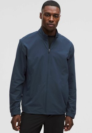 Blaue leichte Jacke mit hohem Kragen, durchgehendem Reißverschluss und elastischen Bündchen. Das Material wirkt glatt mit matter Oberfläche.