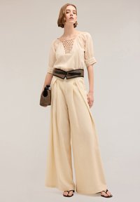 Beige blouse met gestructureerd brei-detail, gecombineerd met brede crèmekleurige broeken. Accessoires zijn een zwarte riem en een geweven tas.