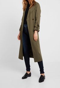 Abrigo de trinchera verde oliva con botones de doble botonadura, cuello con muesca y mangas remangadas, combinado con jeans oscuros y mocasines negros.