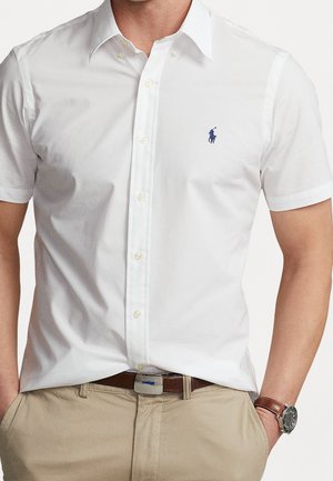 Korte mouwen, wit katoenen shirt met een kraag, voorzien van een klein blauw geborduurd logo. Gecombineerd met een beige broek en een bruine riem.
