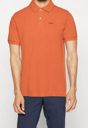 Homme portant un polo orange à manches courtes avec un logo noir "GANT" assorti d'un pantalon bleu foncé, mains détendues le long du corps.