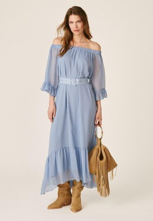 Mujer con vestido azul claro sin hombros con cinturón, botas de ante marrón claro, sosteniendo un bolso marrón claro con flecos y asas redondas, de pie frente a un fondo liso.