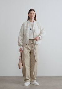 Μπουφάν bomber κρεμ με ραφές, λευκό φούτερ με κείμενο, μπεζ παντελόνι ψηλόμεσο, κρατώντας έναν τσαντάκι ανοιχτό ροζ, λευκά αθλητικά παπούτσια.