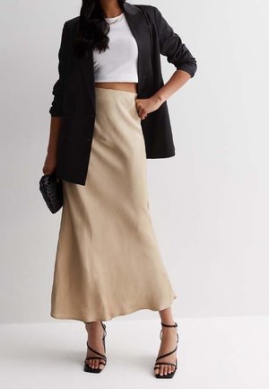 Femme portant un crop top blanc, un blazer noir, une jupe longue beige, des talons noirs à brides et tenant une pochette noire.