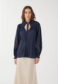 Marinebluse mit hohem Kragen und Bindedetail, mit gerafften Schulterakzenten und langen, leicht puffigen Ärmeln, kombiniert mit einem cremigen Rock.