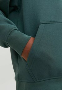 Felpa verde scuro con un ampio taschino frontale, polsini a coste e un tessuto morbido e texture. Il design include cuciture minime e linee pulite.