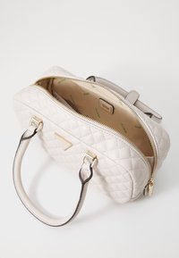 Cremefarvet quilted håndtaske med guldfarvet hardware, lynlås lukning og to top-håndtag. Beigefarvet interiør med "GUESS" mønster i grøn.