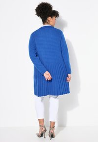 Cárdigan de punto azul con un respaldo texturizado y plisado. Mangas largas y un corte holgado, combinado con pantalones blancos cortos y sandalias plateadas de tacón.