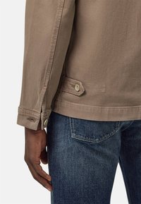 Giacca in tela beige con polsini abbottonati e dettagli sulle tasche laterali, indossata sopra jeans in denim scuro, caratterizzata da una texture liscia e un design classico.