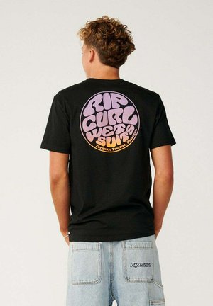 Persoon met krullend haar die een zwart shirt draagt met een rond paars-oranje "Rip Curl Wetsuits" logo en lichtblauwe jeans met "ripcurl" tekst op de zak.