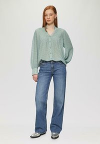 Jasnozielona, zapinana bluzka z teksturowanymi fałdami i ciemnymi kropkami, w zestawie z niebieskimi spodniami z szerokimi nogawkami i metalicznymi srebrnymi butami.