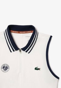 Polo sans manches blanc avec col rayé marine, fermeture éclair, logo Lacoste et patch Roland Garros sur le côté gauche. Texture lisse.