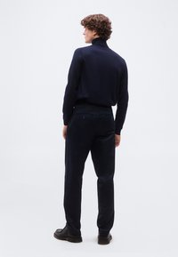 Άντρας με σγουρά μαλλιά στέκεται πλάγια, φοράει μπλε πουλόβερ με λαιμόκοψη, σκούρο παντελόνι και μαύρα παπούτσια μπροστά σε απλό φόντο.