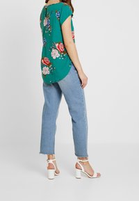 Květinová blůza v zelené barvě, krátké rukávy, s kulatým výstřihem; spárováno se světle modrými džíny a bílými sandály na blokovém podpatku.