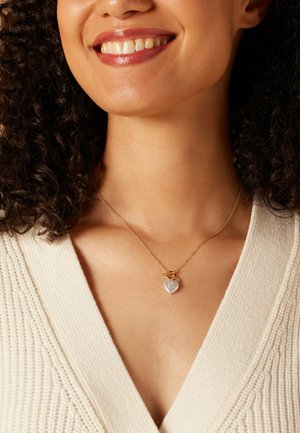 Collier en or avec un pendentif en perle en forme de cœur et un petit charme, porté sur un pull texturé crème clair.
