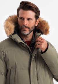 Grüne isolierte Parka mit einer mit Fell gefütterten Kapuze, Reißverschluss vorne und Druckknopfverschluss. Enthält zwei Seitentaschen und ein kariertes Hemd darunter.