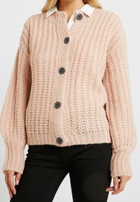 Kvinna iklädd en ljusrosa grovstickad knappradad cardigan över en vit skjorta med krage och svarta byxor, med långa ärmar och ribbade muddkanter.