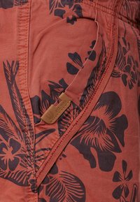 Gros plan d’un short en tissu orange rougeâtre avec imprimé floral noir, poche latérale et une petite étiquette en cuir marron indiquant « EST. 1993. »