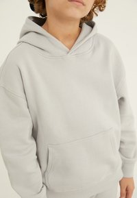 Gaiši pelēks kapucis izgatavots no mīksta kokvilnas ar lielu priekšējo kabatu, regulējamu kapuci un ribotām piedurknēm. Gluda struktūra, ikdienišķs dizains.