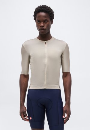 Athletische Person trägt ein beige eng anliegendes kurzärmeliges Fahrradtrikot und marineblaue gepolsterte Fahrradhosen, steht vor einem schlichten Hintergrund.