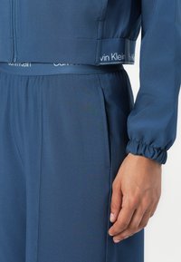 Krátká bunda Calvin Klein v tmavě modré barvě, se zipovým zapínáním, elastickými manžetami a logem na pásku, která zdůrazňuje hladkou texturu látky.