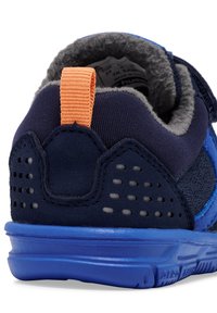 Chaussure de sport bleue avec un dessus marine, une languette orange, une doublure en polaire grise et des détails texturés au niveau du talon. Présente une semelle bleue robuste.