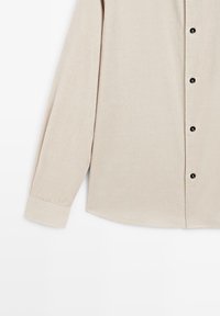 Beige långärmad skjorta i ribbat tyg, med en knappstängning framtill med svarta knappar och en rundad nederkant.
