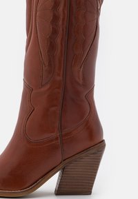 Bota vaquera de cuero marrón con un diseño curvado, costuras decorativas y un tacón de bloque de madera. Textura suave con costuras visibles.