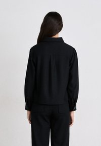 Blazer de mezcla de lana negra con collar estructurado, hombros definidos y puños abotonados. Presenta una silueta limpia y ajustada, con un detalle mínimo.