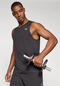 Zwarte mouwloze sporttanktop met een ronde halslijn, voorzien van een klein wit logo op de borst, vastgehouden door een persoon die een zwarte dumbbell vasthoudt.