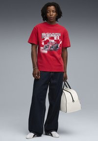 Rød bomulds t-shirt med et Scuderia Ferrari grafisk motiv, parret med løse mørke denim jeans og en hvid taske med sorte detaljer.