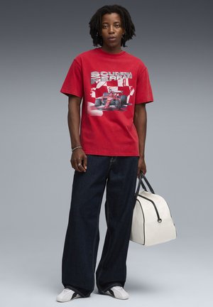 Rød bomulds t-shirt med et Scuderia Ferrari grafisk motiv, parret med løse mørke denim jeans og en hvid taske med sorte detaljer.