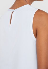 Ärmlös ljusblå topp med en liten knäppning och nyckelhålsdetalj vid övre delen av ryggen, bärs av en person med medelhudton.