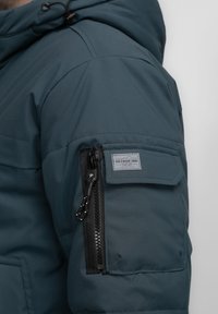 Mørkegrøn pufferjakke med en struktureret hætte, udstyret med en lynlåslomme på ærmet og et logopatch over lommen. Glat tekstur.