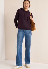 Sudadera morada con cordones ajustables, combinada con unos vaqueros de corte relajado en azul claro. Los accesorios incluyen un bolso marrón de hombro y zapatillas blancas.