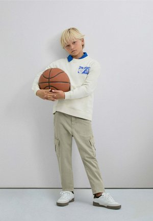 Sudadera en blanco roto con detalles en azul, pantalones cargo color caqui, sosteniendo una pelota de baloncesto, con zapatillas blancas que tienen suelas grises.