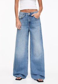 Jean denim bleu à jambes larges avec une taille haute, des poches avant et un léger effet usé. Texture douce, lavage délavé et bords non ourlés.